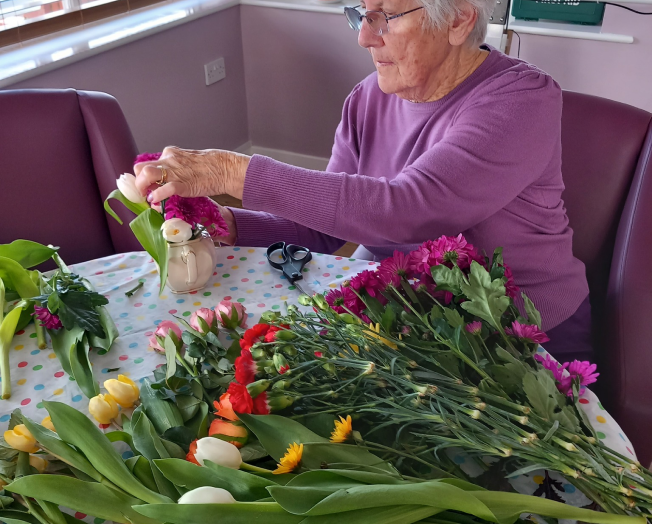 Photo of one of our residents sat at a table which is covered in flower stems, as she is arranging them into a small posy for a jar