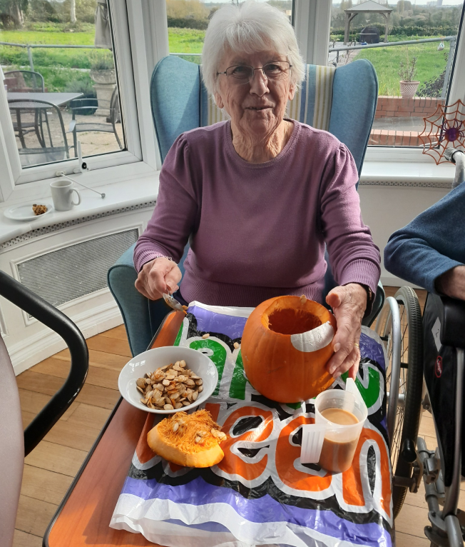 Photo of a resident participating in a Halloween activity. She is sat in an armchair in front of a window with a table in front of her. On the table is a pumpkin which has been emptied into the bowl on the table.
